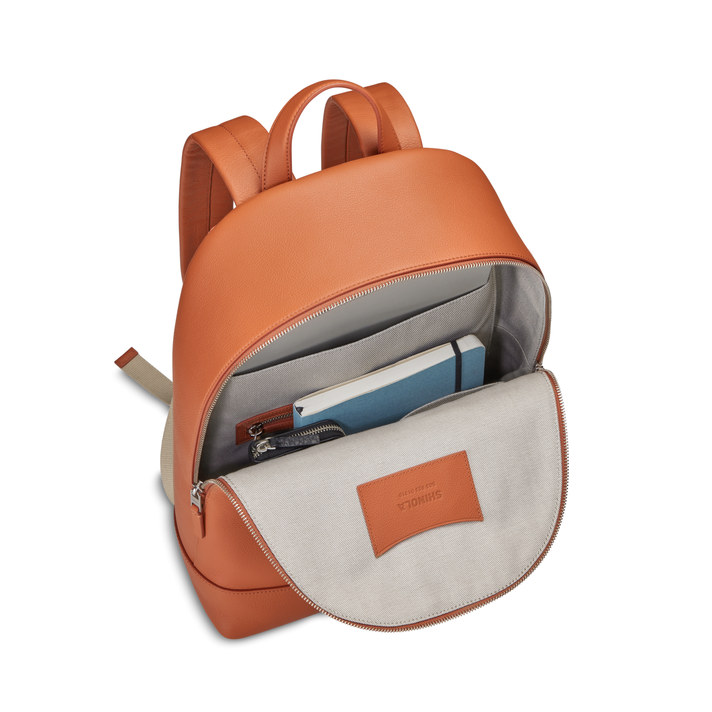 A Shinola Canfield Backpack in rust is unzipped, showing a blue notebook, gray pouch, and white book neatly organized inside its light gray interior.