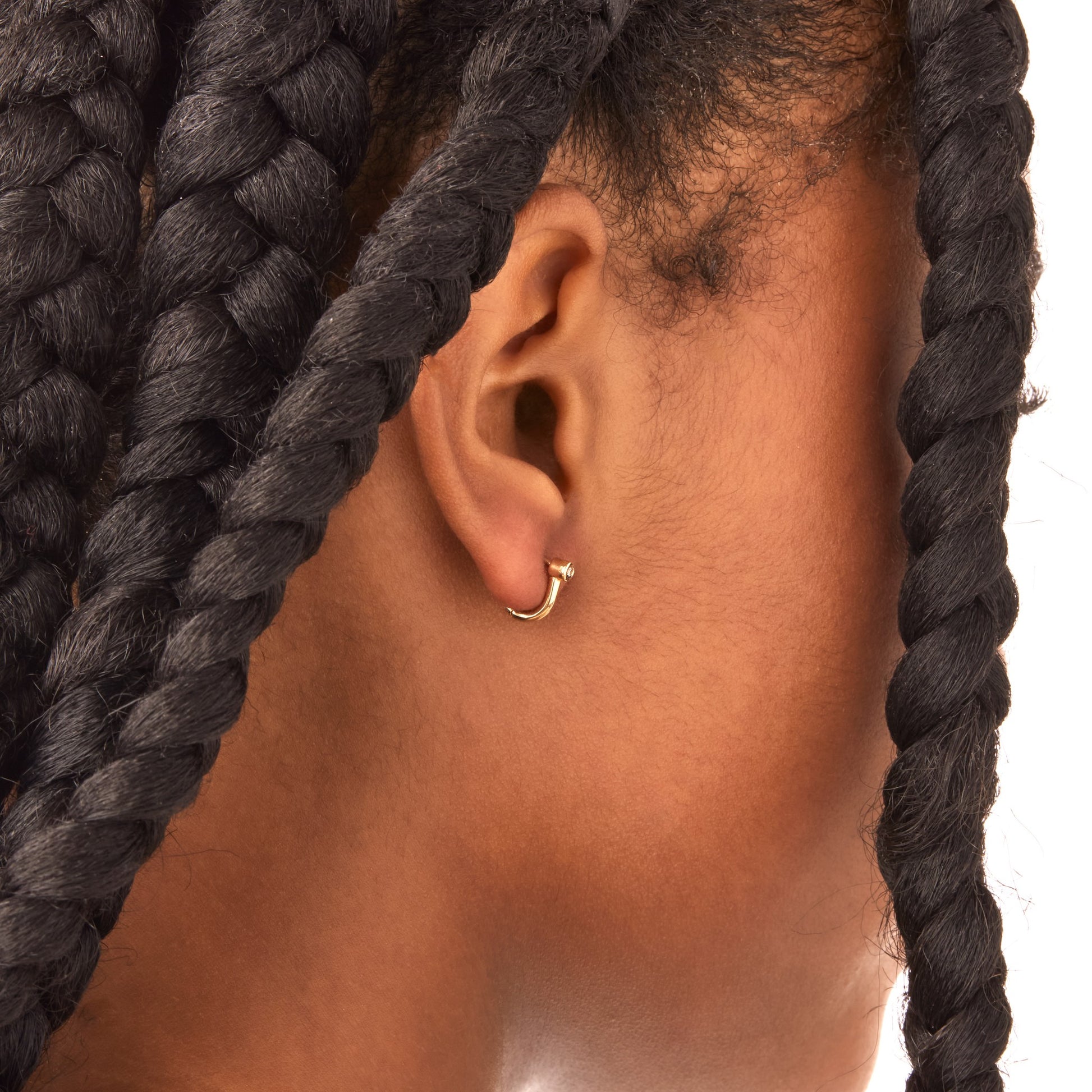A close-up of a person with dark skin and thick braided hair showcases Shinola's Diamond Bike Lock Huggie Earrings in yellow gold, elegantly framing the ear against a plain white background.