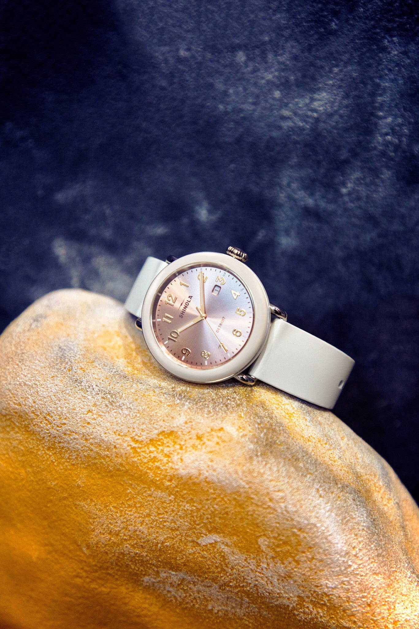 The Shinola Detrola Watch - Nude, showcasing classic American design with a silver case, light gray band, and iridescent face with gold numbers, rests on a textured yellow and gray rock against a dark background.
