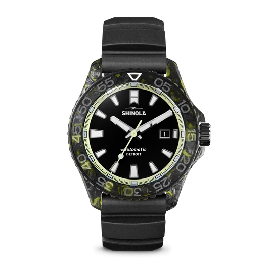 The Shinola Forged Carbon Monster Watch features a black dial with white hour markers, date display, luminous hands, a black and green marbled bezel, forged carbon case, black rubber strap, and Swiss automatic movement.