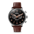 The Shinola Canfield 2 Eye Chrono - Black features a black dial, rose gold hands and numbers, two subdials, date window, brown leather strap, stainless steel silver case, and two buttons on the right side.