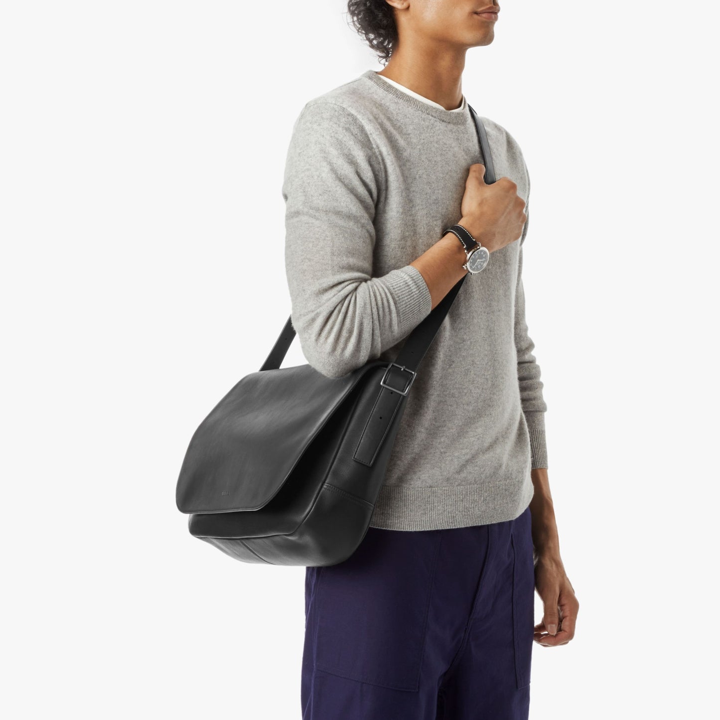 A person in a light gray sweater and dark blue pants carries the Shinola Runwell Messenger Bag - Black with an adjustable crossbody strap over their shoulder against a plain white background.