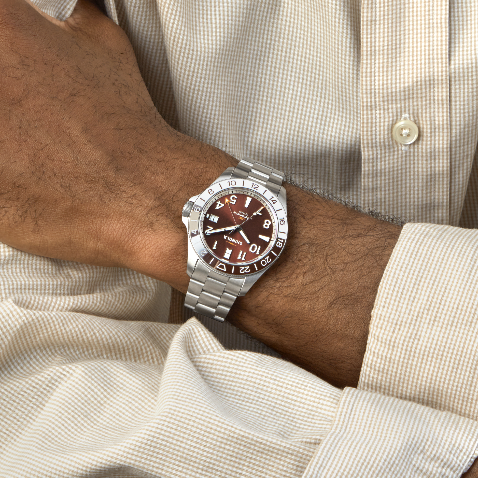 A person in a beige checkered shirt crosses their arms, showcasing a Shinola Monster GMT Automatic Watch – Whiskey with a brown dial and 24-hour bezel on their left wrist.