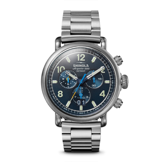 The Shinola Runwell Chronograph West Wind Watch Gift Set features a 42mm silver stainless steel case, blue luminous dial with three subdials, white numerals, Argonite-5050 movement, branding, bracelet strap, and extra strap included.