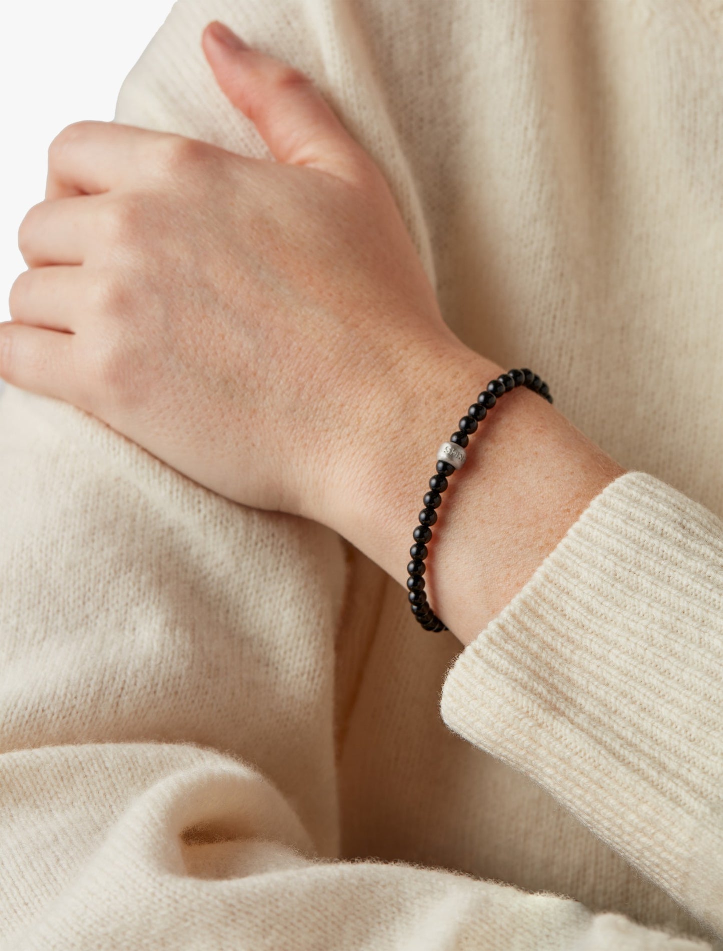 A person in a cream sweater rests their hand on their wrist, showcasing the Shinola 4mm Beaded Onyx Bracelet—an elegant onyx accessory with a single sterling silver accent.