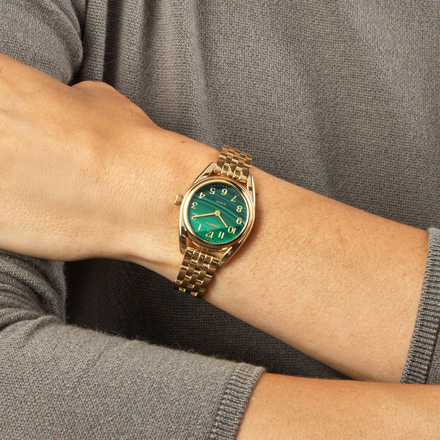 A person in a gray long-sleeve top wears the Shinola Derby Watch - Malachite, crafted in Detroit with a gold stainless steel case, malachite dial, gold numbers, and a uniquely tilted face design.