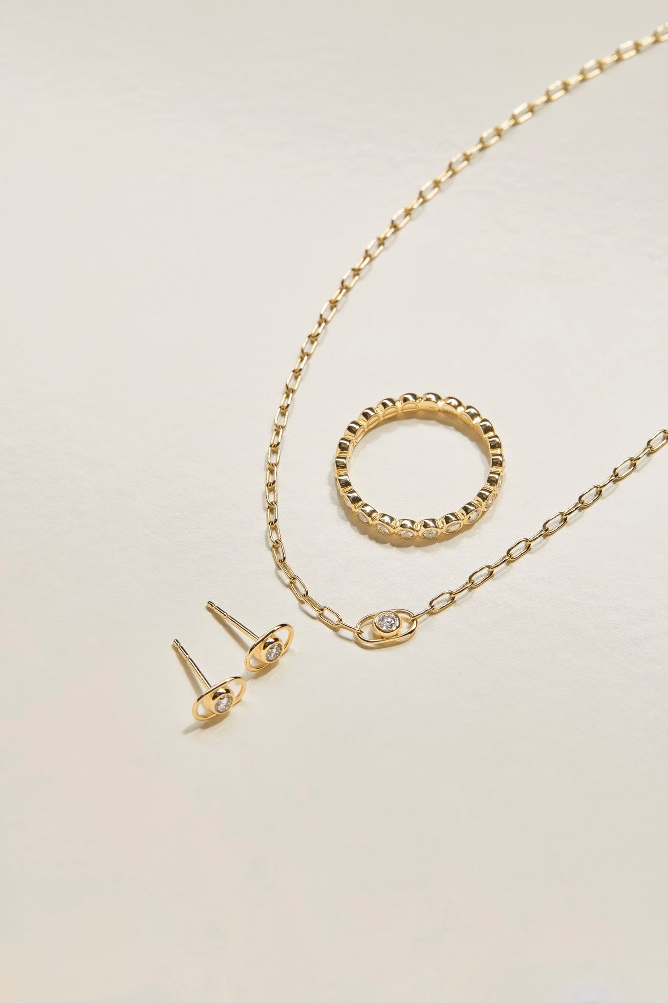 The Shinola Diamond Rivet Oval Stud Earrings in 14k yellow gold feature a small clear stone, shown on a light, neutral background alongside a beaded ring and an eye-shaped pendant chain necklace.