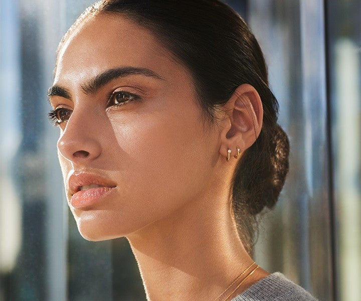 A woman with dark hair pulled back gazes thoughtfully in sunlight, wearing Shinola’s Diamond Bike Lock Huggie Earrings in yellow gold. Her neutral makeup and the softly blurred background add to the serene mood.