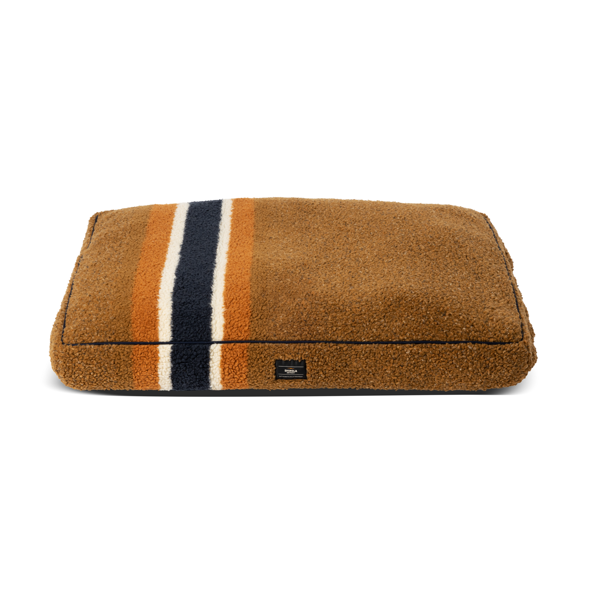The Shinola Napper Pillow Dog Bed - Stripe Brown features orange, cream, and dark blue lengthwise stripes, a soft plush texture, memory fiber fill for comfort, a small black front label, and a machine washable cover for easy cleaning.