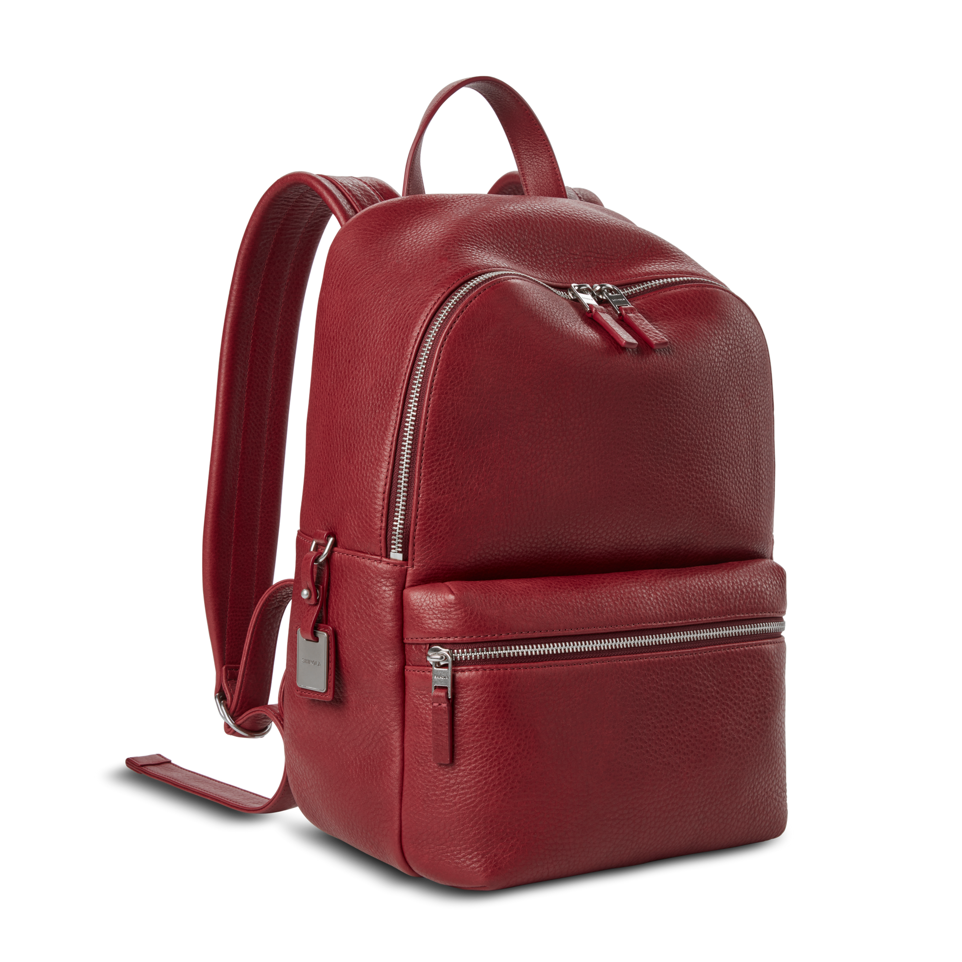 The Shinola Small Runwell Backpack - Ruby is a compact backpack in red Natural Grain Leather, with a front zip pocket, silver zippers, side luggage tag, and adjustable straps, shown on a plain white background.