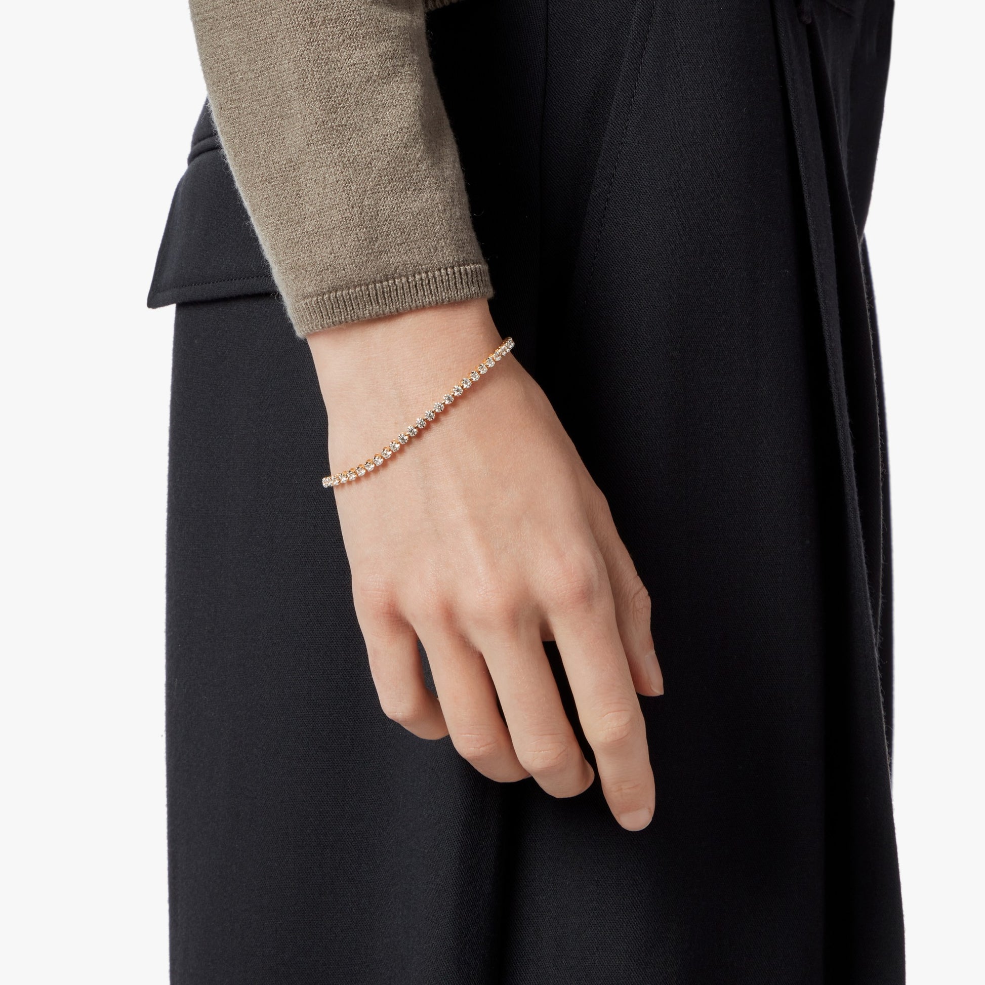 A person in a beige long-sleeve top and dark pants wears the Shinola 1.84 Carat 6.5" Diamond Tennis Bracelet in yellow gold on their wrist against a white background.