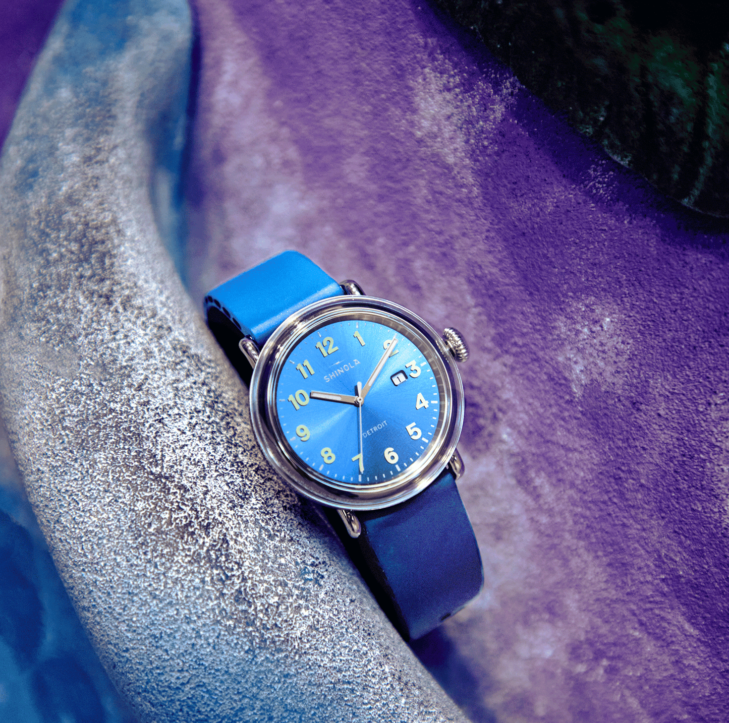 The Shinola Detrola Watch - Denim Blue, featuring a blue face with yellow numbers and a matching strap, rests on a textured, curved stone surface in purple and gray. This American-designed watch displays the date in a sleek silver case.