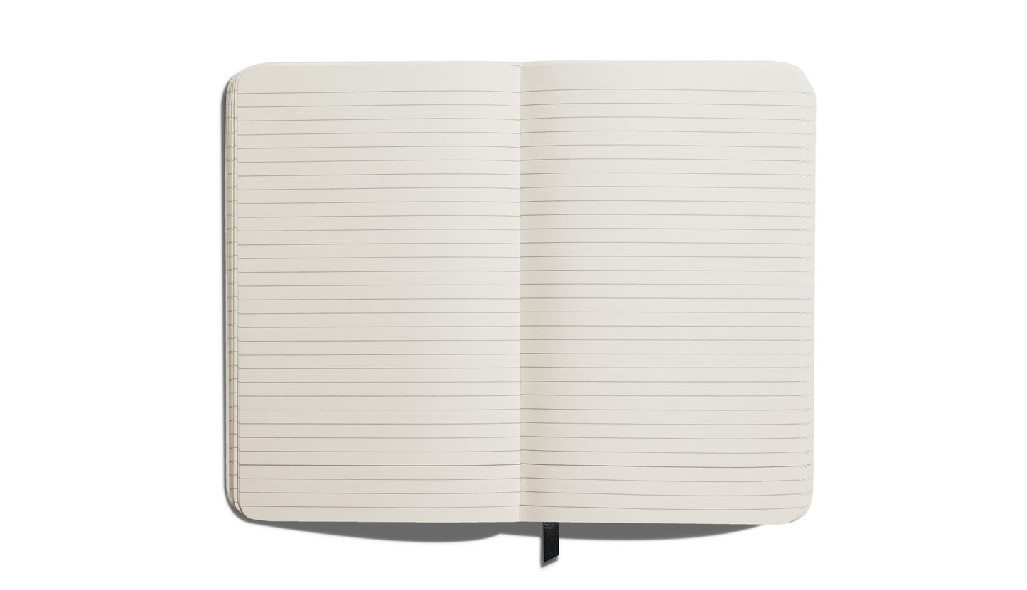 The Shinola Medium Hard Ruled Journal in black features an open, blank, lined interior with rounded corners, a black ribbon bookmark, and smyth-sewn binding, photographed from above on a white background.