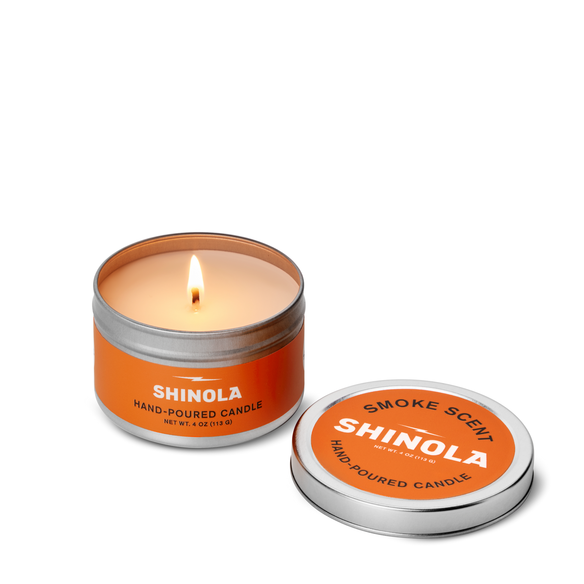 A lit Shinola Hand Poured 4oz Travel Candle - Smoke rests in an orange tin labeled SHINOLA, crafted in Detroit, with a removable lid beside it displaying "Smoke Scent SHINOLA" on a white background.