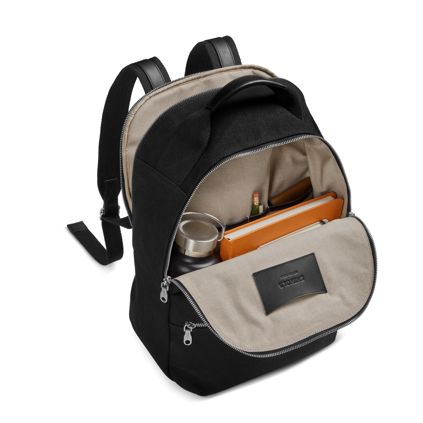 The Shinola Runwell Canvas Backpack - Black features a beige interior and opens to reveal a notebook, metal water bottle, pencils, and small items organized in the main and front compartments.