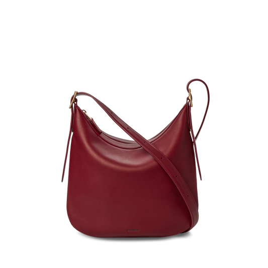 The Shinola Medium Bixby Crossbody - Ruby features natural leather in a curved, crescent shape with gold-tone hardware and an adjustable strap. This versatile shoulder bag is shown against a plain white background.